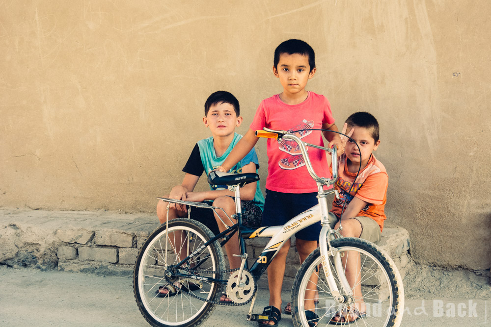 3 Jungs sitzen an der Straßenseite, einer mit Fahrrad. Samarkand, Usbekistan.